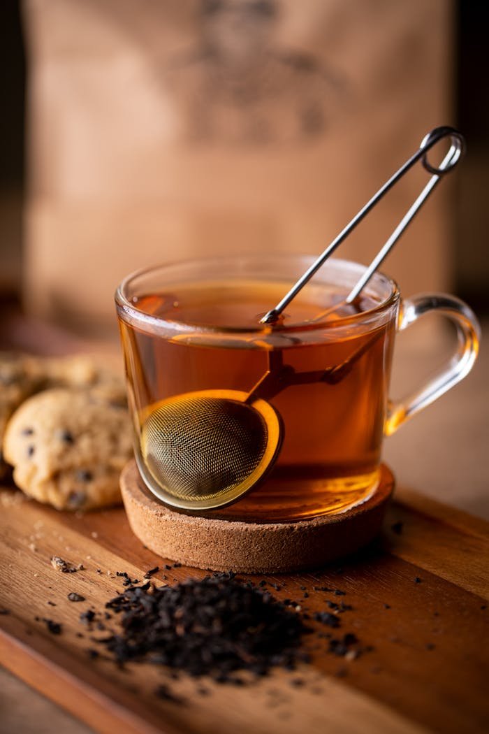 Warm glass cup of freshly brewed tea with a tea infuser and cookies on a wooden surface.